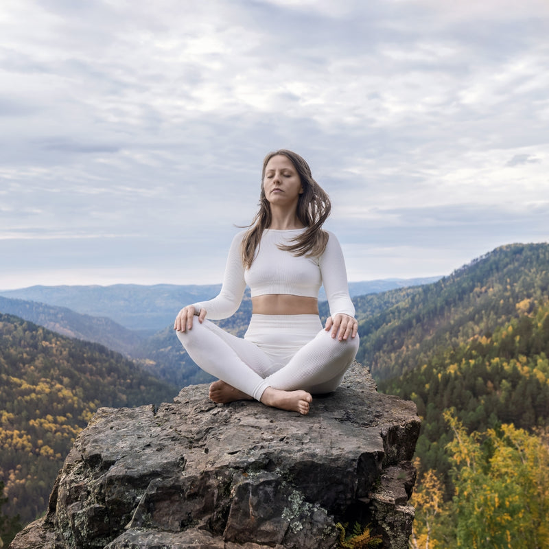 Woman in white outfit meditating on a rocky outcrop with a scenic background